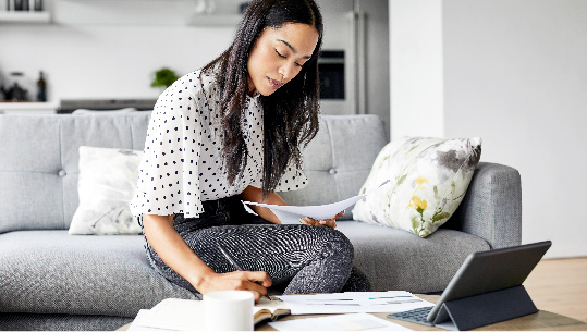 A woman sits on her couch as she writes in her notebook that sits in front of her laptop and a scattering of papers.